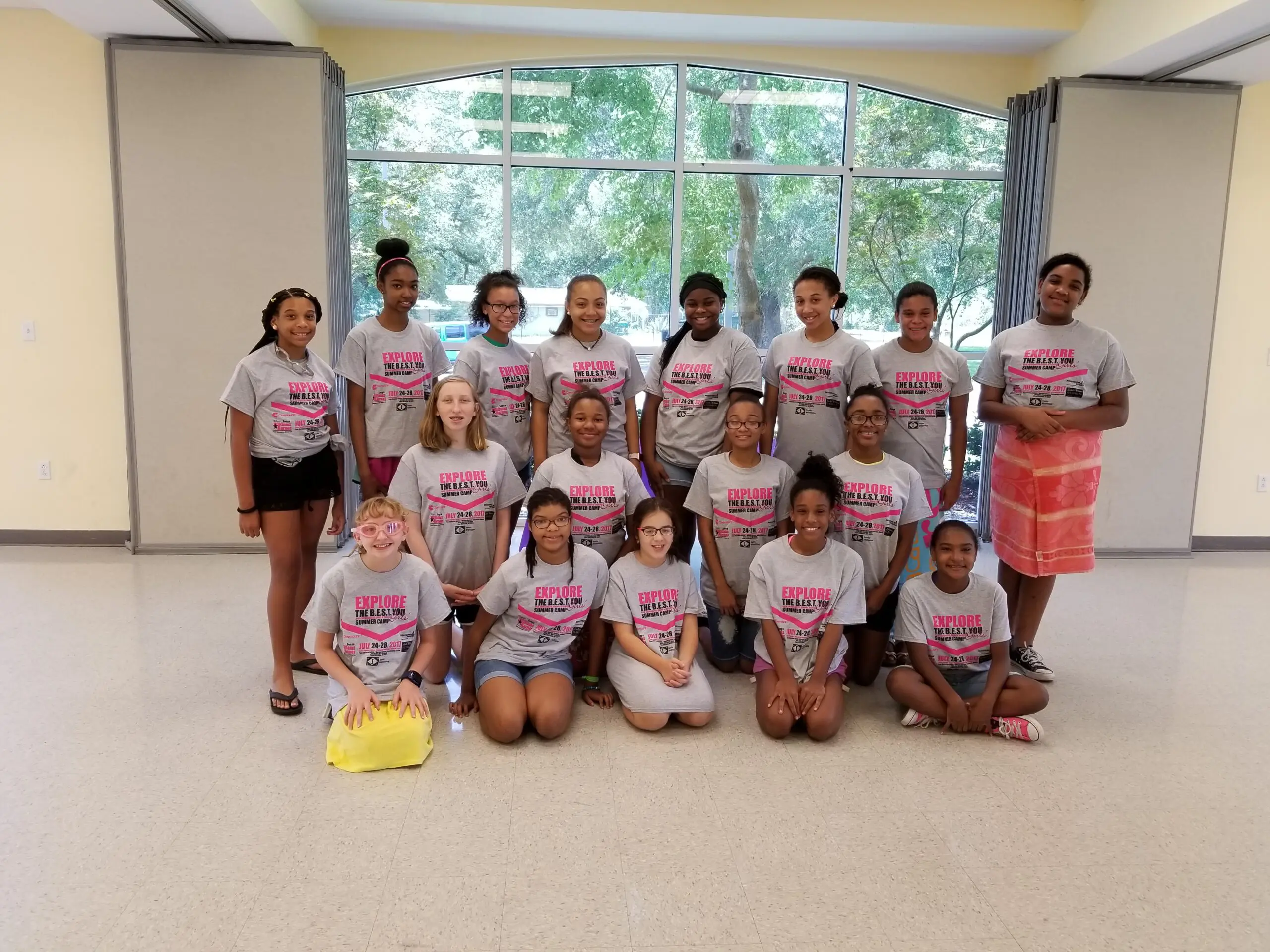 A diverse group of smiling teenage girls posing in gray 'EXPLORE THE B.E.S.T. YOU Summer Camp Girls' t-shirts from July 24-28, 2017, in a brightly lit room with large windows.