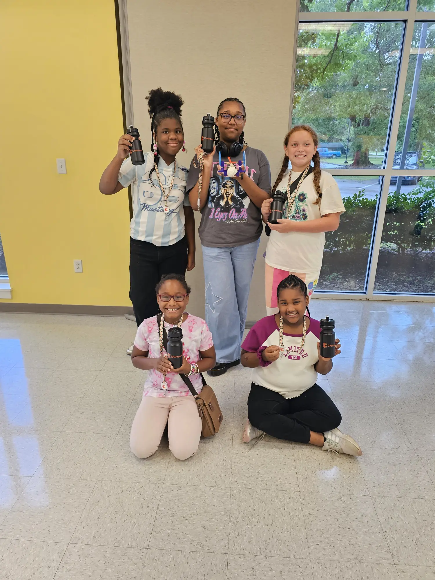 Five diverse children happily showcasing their Chemours water bottles in a bright classroom setting, promoting hydration and community engagement.